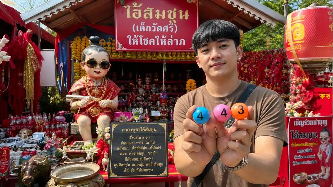 หนุ่มถือบอลตัวเลข 4-4-3 หน้า shrine ไอ้ส้มฉุน ที่วัดทรงเสวย พร้อมของแก้บนสีแดงรอบศาลา ในบรรยากาศกลางวัน